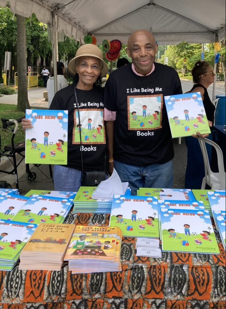 Sam and Rita Burke smiling behind a table of their I Like Being Me books at an outdoor book fair