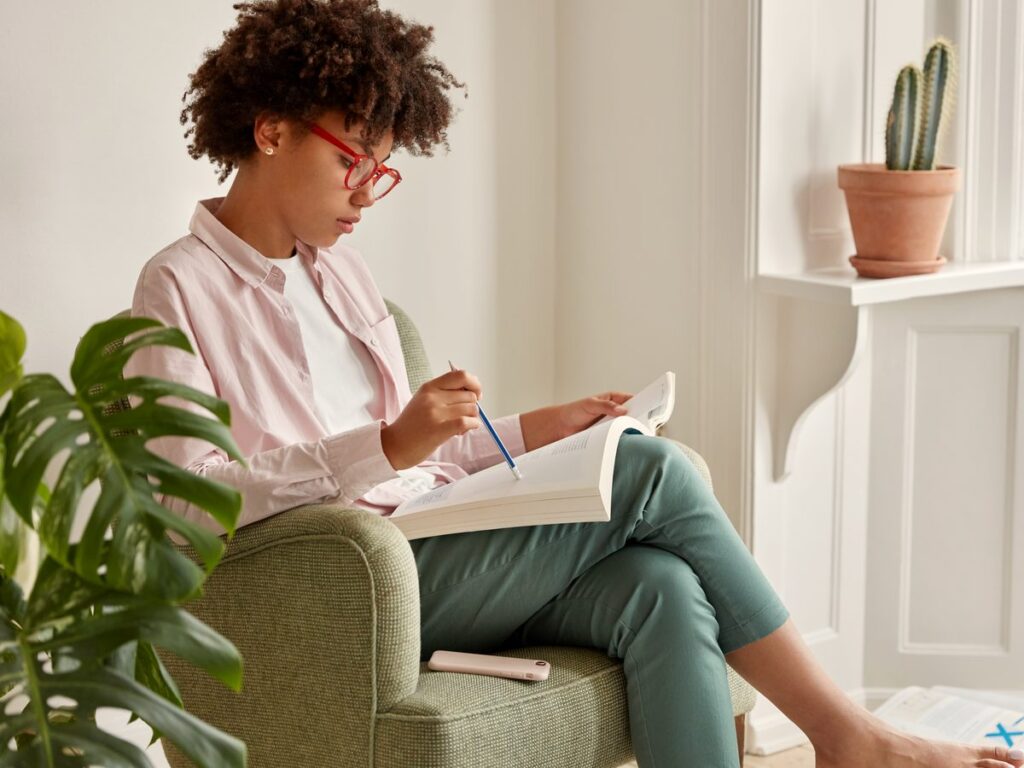 A woman in red glasses reads and makes notes in a book, relaxing in an armchair at home