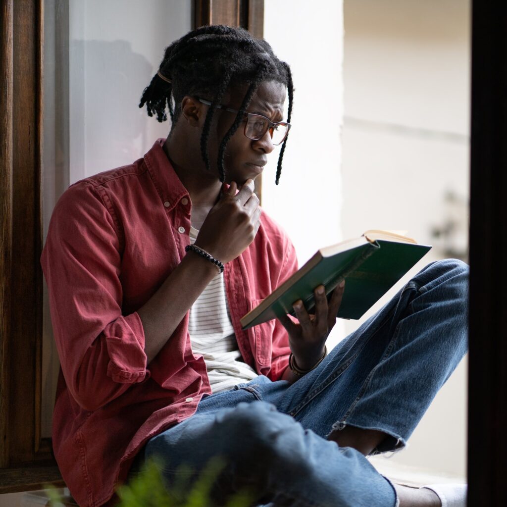 Young Black man in glasses reading a book on a sunny windowsill