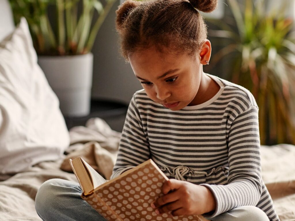A young girl in a striped top reads a picture book, sitting cross-legged on her bed